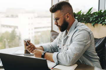 Hipster man sits in cafe, uses smartphone, works on laptop. Businessman reads an information message in phone.