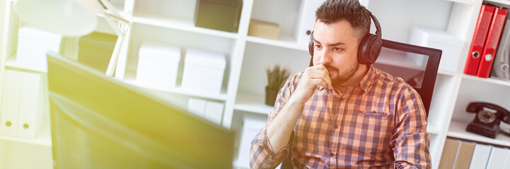 Wall Mural - A young man in headphones sits at a table in the office and looks at the monitor.