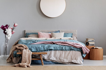 Wall Mural - Trendy round mirror on grey wall in chic bedroom interior with cozy bed with blue, pastel pink and beige sheets and wooden nightstands with books