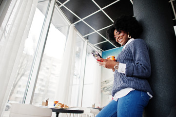 Curly hair african american woman wear on sweater posed at cafe indoor with cup of tea or coffee and mobile phone at hands.