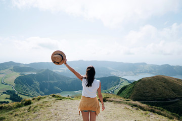 Woman traveler hipster with backpack holding hat and looking forward at amazing mountains and valley view. Azores, Space for text. Wearing stylish boho fall outfit.