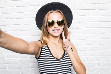 Young woman in straw hat and sunglasses taking selfie smart phone with peace gesture over white brick office wall