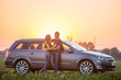 © bilanol - Young couple, slim attractive woman with long hair and handsome sportive man stand embraced together at silver car on warm summer evening on bright sky at sunset or sunrise copy space background.