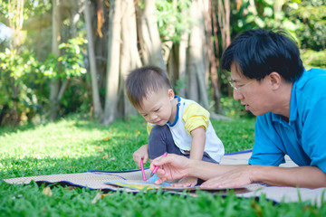 Cute little Asian 2 - 3  years old toddler boy child painting with crayons, kid coloring with father at park, Dad lying on grass spending quality time with son, Creative play for toddlers concept