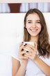 © dianagrytsku - Young smiling attractive brunette woman lies in a big white bed and drinks coffee