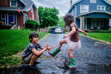 Side View Of Playful Sister Splashing Puddle On Brother During Rainy Season
