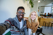 Smiling Young Blond Woman With African American Man Making Selfie While Looking At Camera, Drinking Coffee In Modern Interior Cafe On Background.