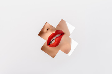 Wall Mural - cropped shot of girl biting red lip through cross shaped hole on white