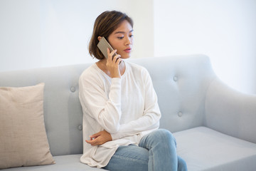 Wall Mural - Portrait of confident young woman talking on cellphone at home. Asian girl sitting on coach and calling up her boyfriend. Wireless communication concept