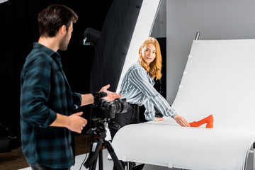 Wall Mural - male photographer standing with camera and looking at colleague arranging shoe in studio