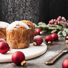 Sprinkling Powdered Sugar Over Traditional Christmas Mini Panettone With Raisins And Dried Fruits On White Marble Serving Plate Surrounded By Red Baubles On Concrete Table Over Black Wooden Background