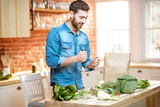 Fototapeta  - Happy man in blue shirt with excited emotions standing near the table full of green fresh vegetables on the kitchen