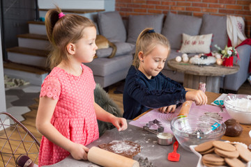 Wall Mural - Young girls baking gingerbread cookies together, rolling dough and making shapes with cookie cutters. Lifestyle, candid image in domestic kitchen