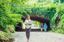 Young East Indian American Woman Traveling, Working In New York, Wearing White Shirt, Black Pants, Holding Laptop Computer, Walking At Central Park. Street Bridge With Green Leaves On Background..