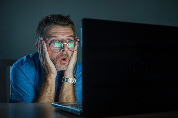 weird shocked and surprised geek man in nerd glasses working on laptop computer late night in the dark looking funny in shock and disbelief face expression