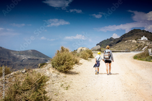 Fotografia, Obraz Mother with son walking on dirt road leading to mountain; Santorini island; Gree