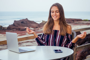 beautiful and calm business woman sitting at a table with a laptop in a summer cafe in the lotus position meditating and relaxing.freelance girl remote work tropics beach paradise island rocks waves