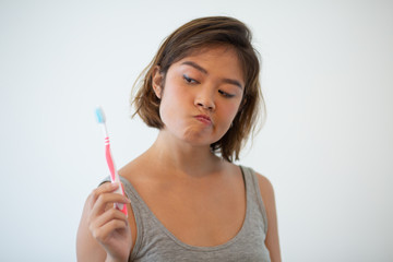 Wall Mural - Doubtful pretty woman looking at toothbrush. Young Asian woman holding tooth brush. Teeth hygiene concept. Isolated front view on white background.