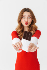 Wall Mural - Portrait of excited woman 20s wearing Santa Claus red costume smiling and eating chocolate bar, isolated over white background