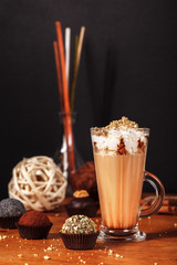 Wall Mural - Beautifully designed latte macchiato coffee with whipped cream and sprinkled with crushed halva, in high transparent glass, on a wooden table, on a dark background with cakes