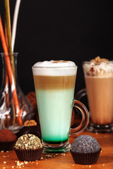 Wall Mural - Coffee latte macchiato with mint syrup and whipped cream, in high transparent glasses, on a wooden table, on a dark background with cakes