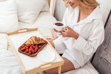 partial view of smiling girl holding coffee cup while sitting on bed with breakfast on tray