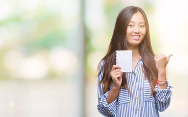 Sticker - Young asian woman holding notebook over isolated background pointing and showing with thumb up to the side with happy face smiling