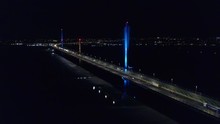 Aerial Flight Around Mersey Gateway Transport Toll Bridge Illuminated Against Night Sky & City Lights On Horizon.