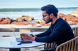 © yurakrasil - freelancing and remote work.handsome male student working laptop keyboard sitting in cafe on the beach with free internet.Young indian man using computer in summer vacation by the sea