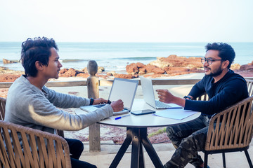 two happy businessmen discussing freelancer strategy in a meeting, Indian business man remote work freelancing sitting in a summer restaurant by the sea.successful deal on the beach tropical island