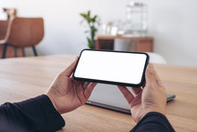 Mockup Image Of Hands Holding Black Mobile Phone With Blank White Screen Horizontally For Watching With Laptop On Table In Office
