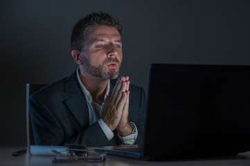 young excited and attractive businessman in praying hands hoping to win online bet while working late night at office laptop computer desk in internet gambling