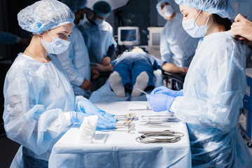 Poster - Nurse s hands holding surgical instruments and tools, close up including scalpels, forceps and tweezers arranged on a table. Surgeons at work in operating room on background.