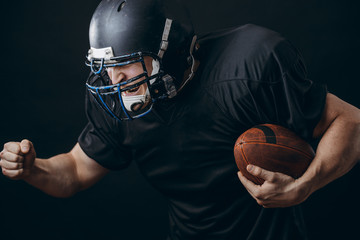 Wall Mural - American football caucasian player wearing black uniform with protection holding ball isolated over black studio wall