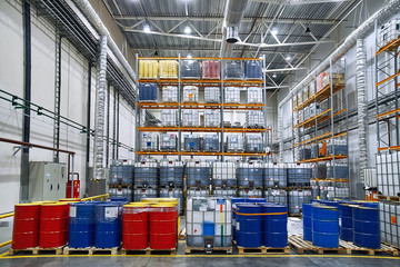 Oil drums and plastic container on pallets in a warehouse on metal shelving. Handling and storing industrial lubricants. Hazardous material storage. Red and blue tank