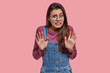 © wayhome.studio  - I think to refuse. Displeased Caucasian girlfriend with straight hair, shows rejection gesture, palms, demonstrates no answer, wears fashionable denim overalls, isolated over pink background