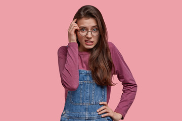 Wall Mural - Indoor shot of pleasant looking young woman gazes in bewilderment, keeps one hand on head, other on waist, wears denim overalls, poses over pink studio wall. Negative facial expressions concept