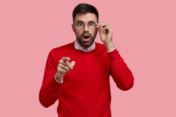 Wall Mural - Waist up shot of stupefied unshaven man points directly at camera with index finger, has scared expression, dressed in red jumper, keeps hand on rim of spectacles, isolated on pink studio wall