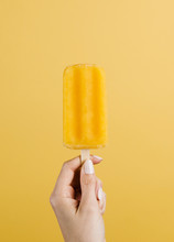 Hand Holding An Orange Popsicle Against A Yellow Background