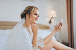 © shchus - Beautiful bride sits on a white bed and looks in a pocket mirror. Wedding interior. Charges the bride at the hotel. Wedding photography. Portrait of a cute bride.