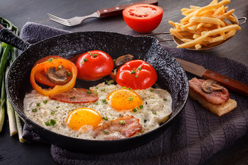 Fried eggs with bacon and tomatoes on an old cast-iron pan with french fries on a gray table. Close-up