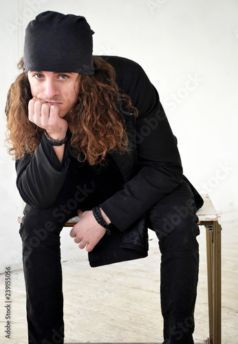 Photo Guy With Long Curly Hair In Black Clothes And A Hat Sits A