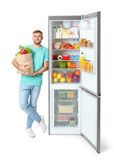 Wall Mural - Young man with bag of groceries near open refrigerator on white background