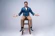 © opolja - Full length portrait of an attractive young man in jeans shirt sitting on the chair over grey background.