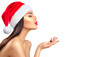 © Subbotina Anna - Christmas winter fashion girl isolated on white background. Beautiful New Year and Xmas holiday makeup. Beauty model woman in Santa's hat blowing snow. Pointing hand, proposing product