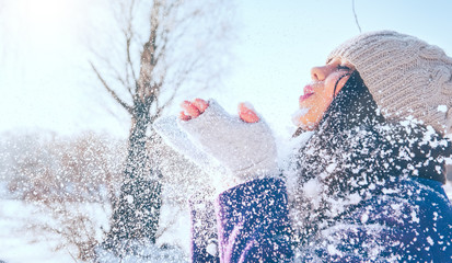 Winter girl portrait. Beauty joyful model girl blowing snow, having fun in winter park. Beautiful young woman enjoying nature outdoors. Wintertime