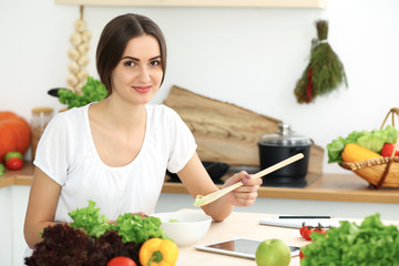 Beautiful Hispanic woman cooking in kitchen while using tablet computer and wooden spoon. Housewife found new recipe for dinner or breakfast. Healthy meal and householding concepts