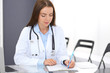 © rogerphoto - Doctor woman at work. Portrait of female physician filling up medical form while sitting at the glass desk at clinic or hospital. Medicine and healthcare concept