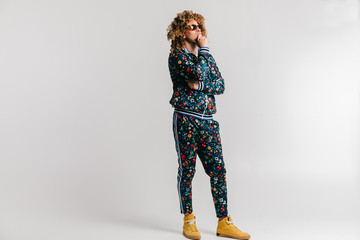 Wall Mural - Adult shylish man in fashionable vintage style tracksuit and sneakers posing on white background. Funky man stroking  beard. Awesome confident male in sunglasses portrait. 80s fashion. Serious person.