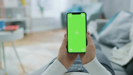 Young Woman at Home Laying on a Couch using with Green Mock-up Screen Smartphone in Vertical Mode.. Girl Using Mobile Phone, Browsing Internet, Videos, Blogs. POV. Screen Has Tracking Markers.